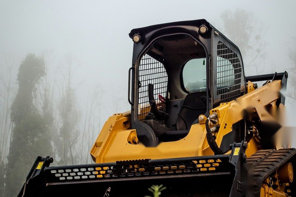 A compact track loader parked at the edge of a field beside a tree line